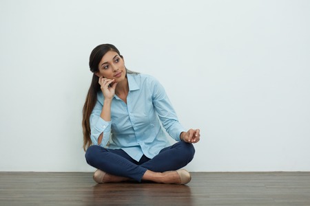 Portrait of pensive young woman sitting on floor with crossed legs, looking aside and leaning on hand with white wall in background. Front view.の写真素材