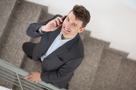 Above view of cheerful Caucasian businessman smiling at camera and talking on phone. Mobile manager having conversation with business partner or client. He standing on stairs. Business conceptの写真素材