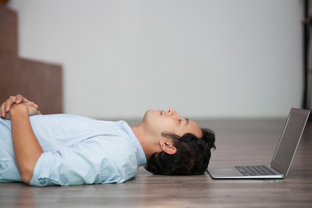 Young Asian man in blue shirt lying on floor at home and looking up with relaxed expression, his laptop nearbyの写真素材