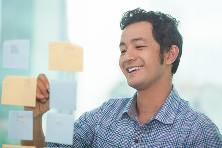 Happy Asian businessman writing down ideas on adhesive notes and smiling happilyの写真素材