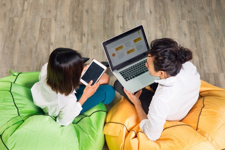 Asian man and woman sitting on bright beanbag chairs in cafÃ© and working on laptop computer and tablet. Top view.の写真素材