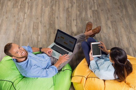 Man and woman sitting on bright beanbag chairs in cafÃ© and working on laptop computer and tablet. Man is turning to camera and smiling. Overhead view.の写真素材
