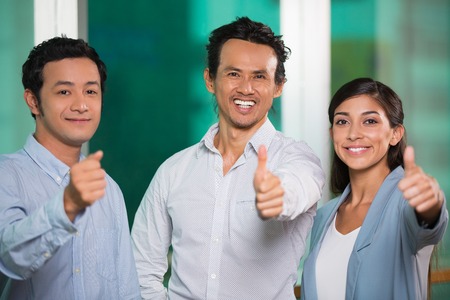 Closeup of three smiling middle-aged business people standing, looking at camera and showing thumbs...