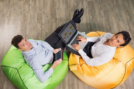 Two serious middle-aged men sitting on bright beanbag chairs in cafÃ©, turning to camera and working on laptop computer and tablet. Top view.の写真素材