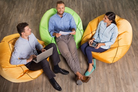 Three smiling young business people sitting on beanbag chairs in office, working and discussing ideas. One man is looking at camera. High angle view.の写真素材