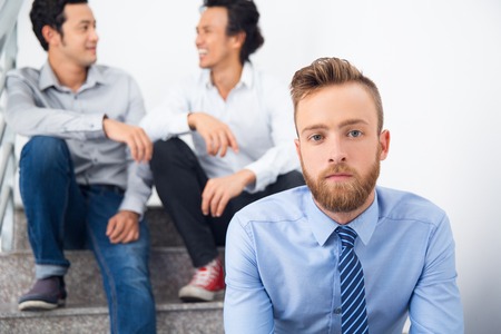 Handsome young businessman sitting on stairs and looking at camera. He resting in corridor during break. His colleagues sitting behind him and talking. Business conceptの写真素材