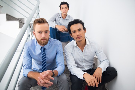 Three serious businessmen sitting on stairs and looking at camera. Young men working as team or they being friends. Multi-ethnic managers resting on stairs. Business team conceptの写真素材