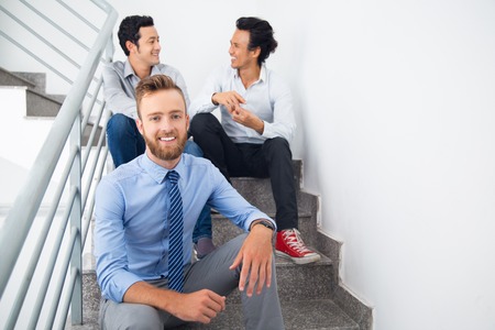 Cheerful young businessman sitting on stairs and smiling at camera. Happy manager and his male colleagues resting and talking in corridor. Business conceptの写真素材