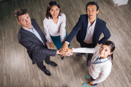 Four smiling adult business people standing in office hall, looking at camera and joining hands together. High angle view.の写真素材