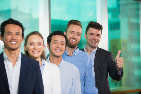 Closeup of five smiling adult business people standing in row and looking at camera. Man in background is showing thumb up. People in foreground are blurred.の写真素材