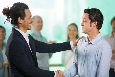 Smiling joyful middle-aged Asian businessmen shaking hands after meeting with four blurred applauding people in background. Side view.の写真素材
