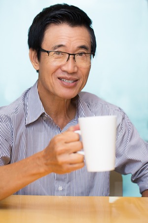 Closeup portrait of joyful Asian senior man looking at camera, sitting at table, holding mug and drinking teaの写真素材