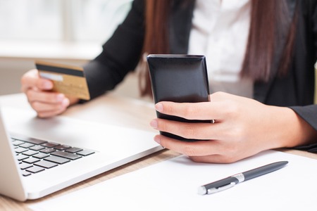Cropped view of business woman working on laptop computer, holding credit card and smartphone and doing online banking at office deskの写真素材