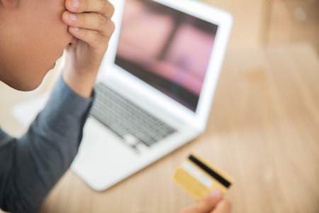 Young Asian businessman sitting at table and holding credit card in office. Manager covering eyes in stress and crying. Debt and loan conceptの写真素材