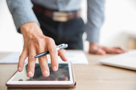 Close-up of hand of unrecognizable businessman standing at table, holding pen and using digital tablet in officeの写真素材