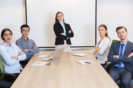 Portrait of confident multiethnic businesspeople sitting at meeting table in boardroom and looking at camera with arms crossed. Young female leader standing in centerの写真素材