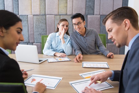 Business team sitting at table at meeting. Young Caucasian man discussing data on paper with young Latin-American woman. Young Caucasian woman looking at them, smiling and whispering to Asian manの写真素材