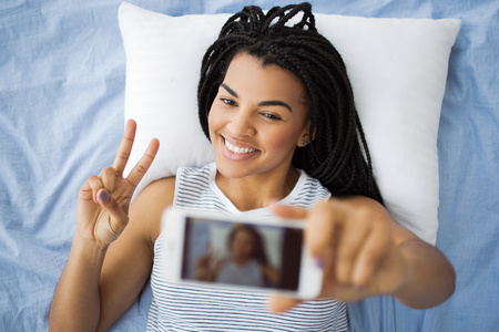 Happy girl taking selfie showing horns sign in bedの写真素材