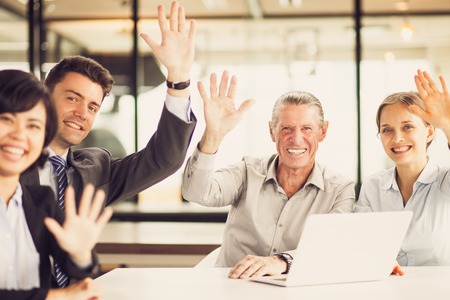 Successful multiethnic business team sitting in boardroom at meeting, waving, looking at camera and smilingの写真素材