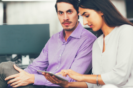 Confident young businesspeople, Latin-American businesswoman and Caucasian businessman, sitting in office and using digital tablet, man looking at cameraの写真素材