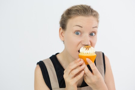 Frowning young woman eating cupcake with greed and looking at camera against white background. Goodbye, diet conceptの写真素材