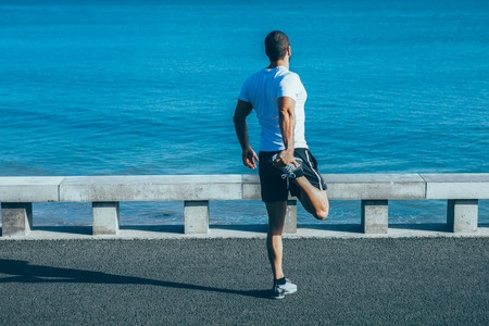 Rear view of athlete stretching leg on road by sea