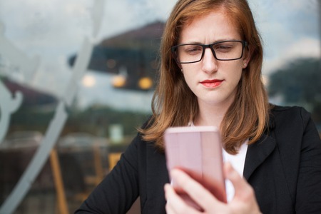 Portrait of displeased young Caucasian woman, businesswoman or student wearing glasses, reading news or message on mobile phone. Bad news, mobile communication conceptの写真素材