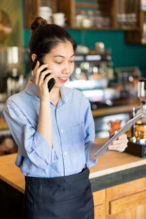 Positive confident Asian manager using mobile phone while talking to supplier and examining check-list on tablet. Small business conceptの写真素材