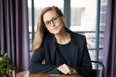 Closeup portrait of confident young beautiful brown-haired woman looking at camera and sitting at cafe table with window in background. Beautiful business woman concept. Front view.の写真素材