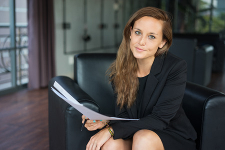 Closeup portrait of content young beautiful brown-haired woman looking at camera, holding papers and sitting in armchair in hotel lobby. Beautiful female company representative concept. Front view.の写真素材