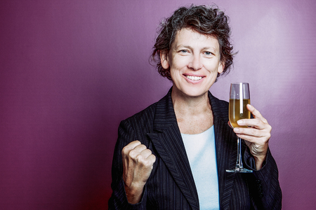 Portrait of successful senior Caucasian businesswoman wearing suit holding champagne flute, looking at camera, showing winning gesture and smiling on purple backgroundの写真素材