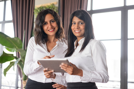 Two happy business ladies using tablet computer in cafe. They are standing and looking at camera with windows in background. Technology concept. Front view.の写真素材