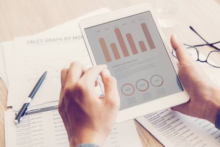 Close-up of hands of businessman sitting at table and holding digital tablet with marketing data on screen. Manager studying information at workplaceの写真素材