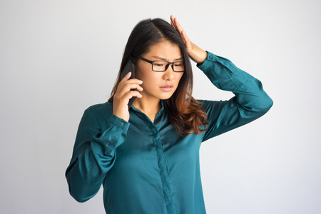 Concerned young Asian woman speaking on phone and touching head. Upset business woman getting bad news. Communication and problem conceptの写真素材