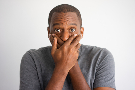 Closeup of shocked African American keeping silence. Frightened black man in grey tshirt covering mouth with hands. Shock and keeping secret conceptの写真素材