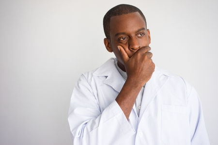 Portrait of upset African man in white coat. Frustrated doctor covering mouth with hand. Bad news and medicine conceptの写真素材
