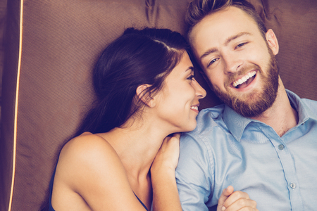 Portrait of smiling young couple enjoying lying together on sofa, man laughing and looking at camera, woman resting on his shoulderの写真素材