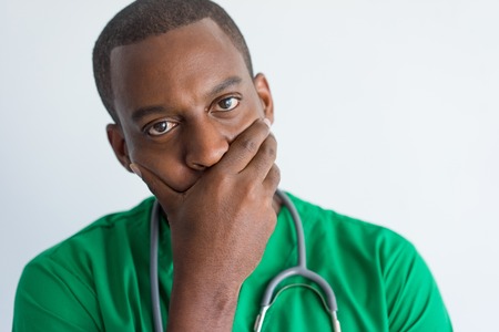 Close-up of pensive young general practitioner covering mouth. Face of African American doctor wearing green medical garment with hand on chin. Trouble conceptの写真素材
