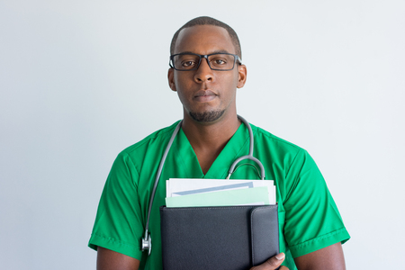 Close-up of serious young African American doctor with folder. Face of general practitioner wearing glasses and green scrubs holding medical records and looking at camera. Medical staff conceptの写真素材