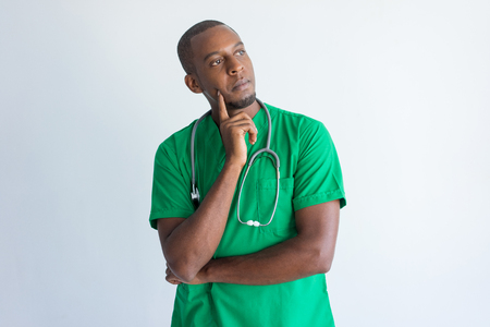 Portrait of contemplated doctor with hand on chin looking away. Young African American general practitioner wearing green medical garment and with stethoscope. Contemplation conceptの写真素材