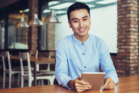 Closeup portrait of smiling young Asian man looking at camera, using tablet and sitting at empty table with blurred cafe interior in backgroundの写真素材