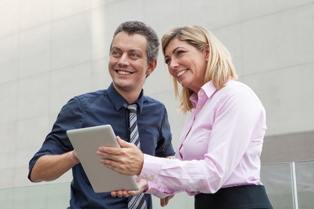 Smiling male and female business people using tablet computer outdoors. Businesspeople standing and discussing issues with building wall in background. Technology in business concept.の写真素材