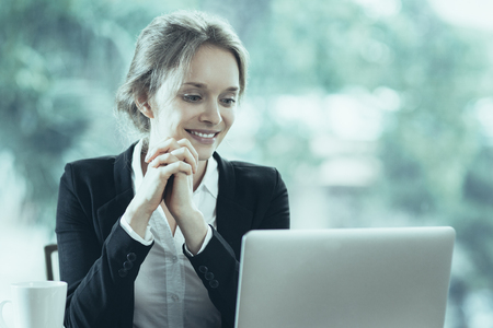 Smiling young attractive businesswoman sitting at table while looking at laptop screen with her hands clasped. Front view with big window and blurry green view outside in background.の写真素材