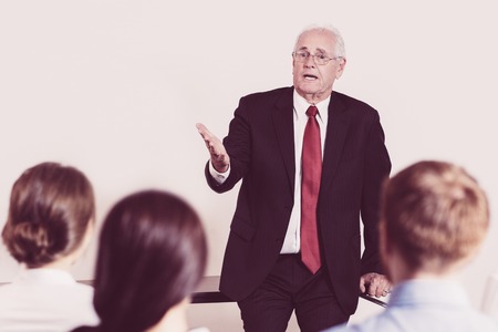 Senior businessman leaning on table, gesturing and telling something to sitting audience of four...