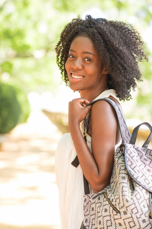 Smiling attractive young black woman holding rucksack, turning round, looking at camera and walking in park with blurred plants in background. Park relaxation and tourism concept.の写真素材