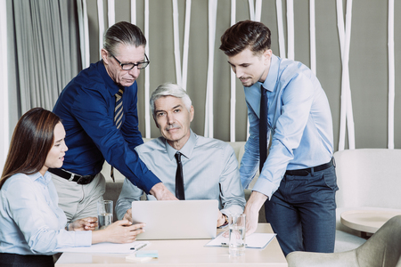 Content young, two senior businessmen and middle-aged businesswoman working and discussing ideas while sitting and standing at table with laptop in cafe. Sitting senior man is looking at camera.の写真素材