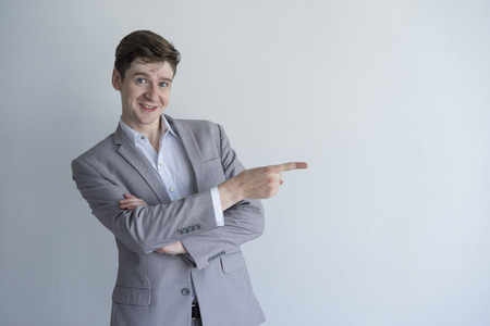 Positive enterprising male manager pointing aside and looking at camera. Cheerful young businessman in gray suit gesturing and standing against isolated background. Presentation conceptの写真素材