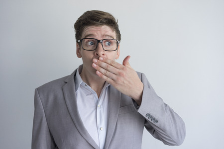 Surprised frowning young businessman in eyeglasses covering mouth and looking away. Shocked man in gray jacket knowing news. Gossip conceptの写真素材