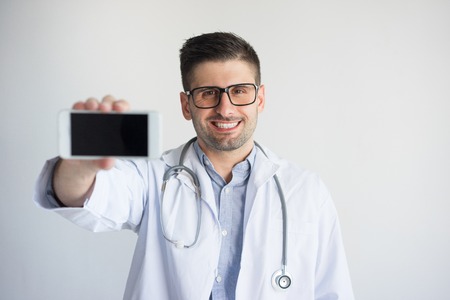 Portrait of happy male general practitioner showing smartphone. Young Caucasian medic wearing glasses, lab coat and stethoscope advertising medical software. Medical app conceptの写真素材