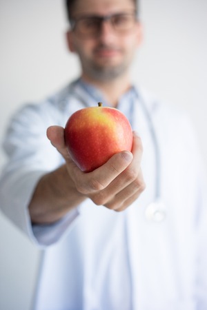 Close-up of doctor hand giving red apple. Young Caucasian male dentist or nutritionist offering apple. Healthy eating and detox dieting conceptの写真素材